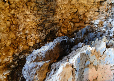 La capilla fue descubierta durante la campaña arqueológica de 2009, en la falda de la colina de Dra Abu el-Naga, a una altura que corresponde con el tercer nivel o piso justo por encima de la tumba-capilla de Hery (TT 11; ubicada a los pies de la colina, en el primer nivel). Accedimos a su interior a través de un gran agujero en el techo que se formó tras ceder el dintel de la entrada. Todavía no está del todo clara la planta completa de la capilla, pues ésta fue posteriormente reutilizada y su estructura alterada en el siglo II a. C., y queda todavía por excavar una sala contigua que está llena casi hasta el techo de tierra y derrubio de la colina. La sala que sí hemos excavado y que ahora está visible tiene unas dimensiones de 2,00 x 2,40 m y una altura de 1,60 m aproximádamente. Las paredes conservan la decoración pintada de la mitad inferior, desde el suelo hasta una altura de unos 0,80 m. Gracias a la inscripción que acompaña a una escena de manipulación de telares y elaboración de telas, sabemos que la capilla perteneció a un “supervisor de tejedores” llamado Ramose, que debió vivir probablemente bajo el reinado de Ramsés II, ca. 1200 a. C. Limpieza y consolidación de las pinturas de la capilla ramésida Limpieza y consolidación de las pinturas de la capilla ramésida Limpieza y consolidación de las pinturas de la capilla ramésida Mientras que a los pies de la colina la buena calidad de la roca caliza permitió a Hery tallar en relieve la decoración de las paredes interiores de su monumento funerario, a la altura del tercer nivel la roca es poco compacta y está totalmente meteorizada, lo que imposibilita la decoración en relieve y obliga a que ésta sea pintada. La pintura está realizada sobre una capa gruesa de preparación, compuesta por dos estratos. El primer estrato, el que contacta con la roca, es grueso, tosco y oscuro, de entre 3 y 4 cm de espesor, y está compuesto básicamente por barro y abundantes fibras vegetales (paja). Su utilidad es dar consistencia a la roca tallada y uniformar la superficie. El segundo estrato, de 1 cm. de espesor aproximadamente, es fino, cuidado y más claro, compuesto posiblemente de yeso, y destinado a recibir la policromía. Limpieza y consolidación de las pinturas de la capilla ramésida Limpieza y consolidación de las pinturas de la capilla ramésida Limpieza y consolidación de las pinturas de la capilla ramésida El paulatino desgaste y desmoronamiento de la roca poco compacta y meteorizada provoca que la capa de preparación pierda su soporte y tienda a caer y a acumularse, junto con polvo, tierras, y fragmentos de la propia roca, en el espacio que se ha ido abriendo entre la capa de preparación y la roca, haciendo de cuña, ensanchando más el espacio y aumentando los riesgos de caída. En la campaña del 2013 se ha acometido la conservación de las pinturas, que ha consistido en la limpieza, consolidación estructural del soporte y la consolidación superficial de las policromías. Lo primero que se hizo fue arriostrar las pinturas con el fin de trabajar con seguridad y evitar posibles desplomes durante los trabajos de consolidación y relleno. Se colocó un puntal telescópico de un lado a otro de la pared principal, y se encajaron tacos de espuma de polietileno con la presión justa para no forzar la pintura hacia el muro. Una vez realizada esta sujeción preventiva, se procedió a succionar con una aspiradora el material acumulado en el hueco existente entre la pintura y la roca. Tanto la roca como el soporte se consolidaron con Primal AC-33 al 20% aplicándolo en manos sucesivas hasta la saturación. Para consolidar las zonas inaccesibles desde la parte superior, se aprovecharon los múltiples agujeros del frente, carentes de policromía, en los que se practicaron pequeños taladros de no más de un centímetro de diámetro, y a través de ellos se realizaron inyecciones con el mismo consolidante. Una vez secas las superficies consolidadas, el vacio resultante se rellenó con mortero sintético “parrot”, mortero ligero y con gran poder adhesivo. Se rellenó con este mortero hasta unos 4-5 cm. del borde superior, completando el espacio hasta el borde con mortero de cal hidráulica, arena y pigmento para integrarlo visualmente con el conjunto. Este mismo tipo y tono de mortero fue empleado en todas las lagunas de la parte frontal del las pinturas. Limpieza y consolidación de las pinturas de la capilla ramésida Limpieza y consolidación de las pinturas de la capilla ramésida Limpieza y consolidación de las pinturas de la capilla ramésida La capa de barro y tierras duras, de poco grosor, se eliminó reblandeciéndola con agua-alcohol y la ayuda mecánica de bisturí. Algunas zonas estaban ennegrecidas por calcinación debido a la acción de altas temperaturas, como consecuencia de haber realizado hogueras en el siglo II a. C. para quemar allí momias de ibis y halcones (en 2009 se halló en el centro de la sala restos de un fuego con momias calcinadas). En las zonas ennegrecidas no se pudo recuperar el color ya que la calcinación era estructural, y únicamente se pudo realizar una limpieza superficial del hollín con hisopos de agua-alcohol. La superficie policromada presentaba pulverulencias y erosiones que era necesario consolidar. Previamente se realizaron pruebas y se observó que los blancos adquirían un tono amarillento que cambiaba completamente el aspecto de las pinturas. Los demás colores, rojos y azules en su gran mayoría, no variaban. Por esta razón, se decidió consolidar toda la superficie a excepción de los blancos de los fondos y de los ropajes. El consolidante empleado fue Paraloid B-72 al 7%, aplicado con pincel en las zonas seleccionadas. Limpieza y consolidación de las pinturas de la capilla ramésida Limpieza y consolidación de las pinturas de la capilla ramésida Limpieza y consolidación de las pinturas de la capilla ramésida Para completar estos trabajos queda pendiente, en la próxima campaña, cambiar la techumbre de madera que instalamos de manera provisional al final de la campaña de 2009, por un cierre que proteja de forma integral las pinturas, que evite más eficazmente la entrada de tierras y agua, y que amortigüe los cambios bruscos de humedad y, sobre todo, de temperatura. A pesar del reducido tamaño de la sala y del delicado estado de conservación de las pinturas que se conservan en la mitad inferior de las paredes, el monumento es de interés sobre todo por la escena de telares que contiene, pues sólo hay otras cuatro capillas en la necrópolis de Tebas que conserven escenas de este tipo. La representación de distintos momentos de la confección de telas de lino, desde la manipulación de los telares por niños desnudos hasta la muestra y prueba de la pieza final delante del supervisor Ramose, permite conocer más detalles sobre esta cotidiana actividad artesanal a la que tanto valor otrogaban los egipcios en época antigua. Limpieza y consolidación de las pinturas de la capilla ramésida Limpieza y consolidación de las pinturas de la capilla ramésida Limpieza y consolidación de las pinturas de la capilla ramésida